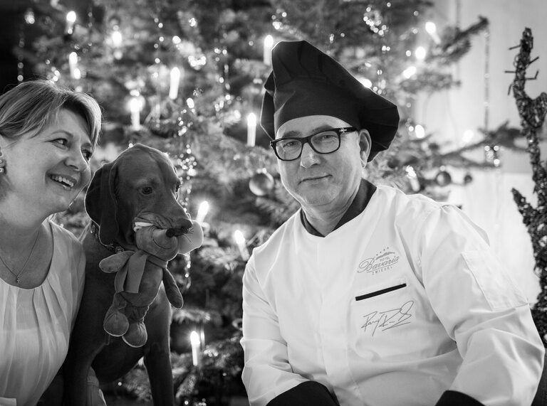 Ein lächelnder Herr im Kochoutfit sitzt mit einer Frau vor einem festlich geschmückten Weihnachtsbaum.